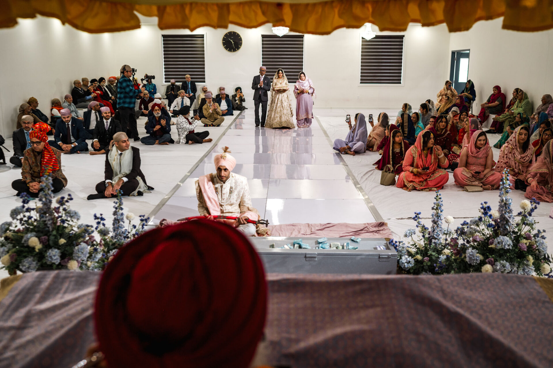 A Sikh wedding ceremony in a hall with seated guests and a couple standing.