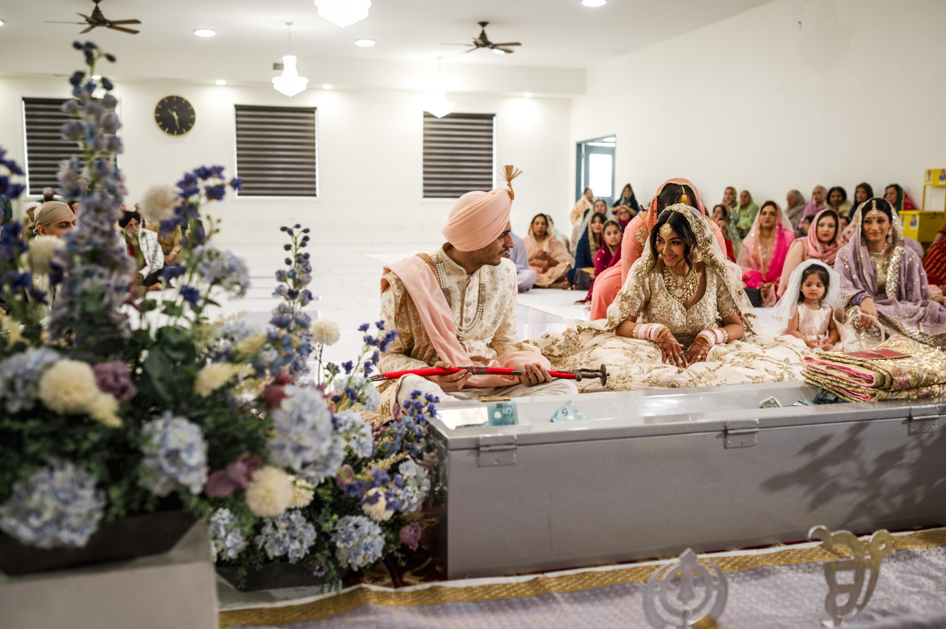 A couple in traditional attire participates in a Sikh ceremony, surrounded by guests and blue flowers.
