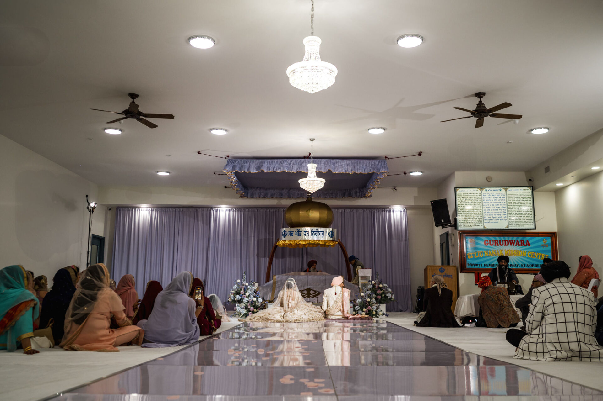 Sikh wedding ceremony in a gurdwara with guests seated, floral decorations, and chandeliers shining.