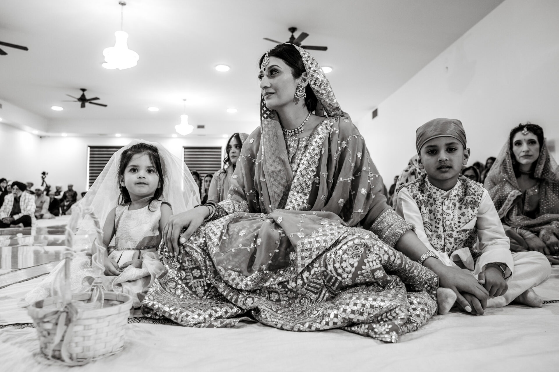 Woman in traditional attire with children at a Sikh event, seated on the floor indoors.