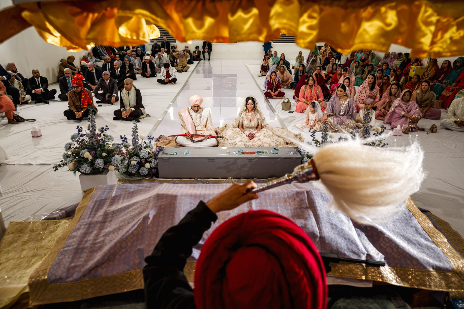 A Sikh wedding: couple seated, guests gathered, seen from behind priest with white whisk.