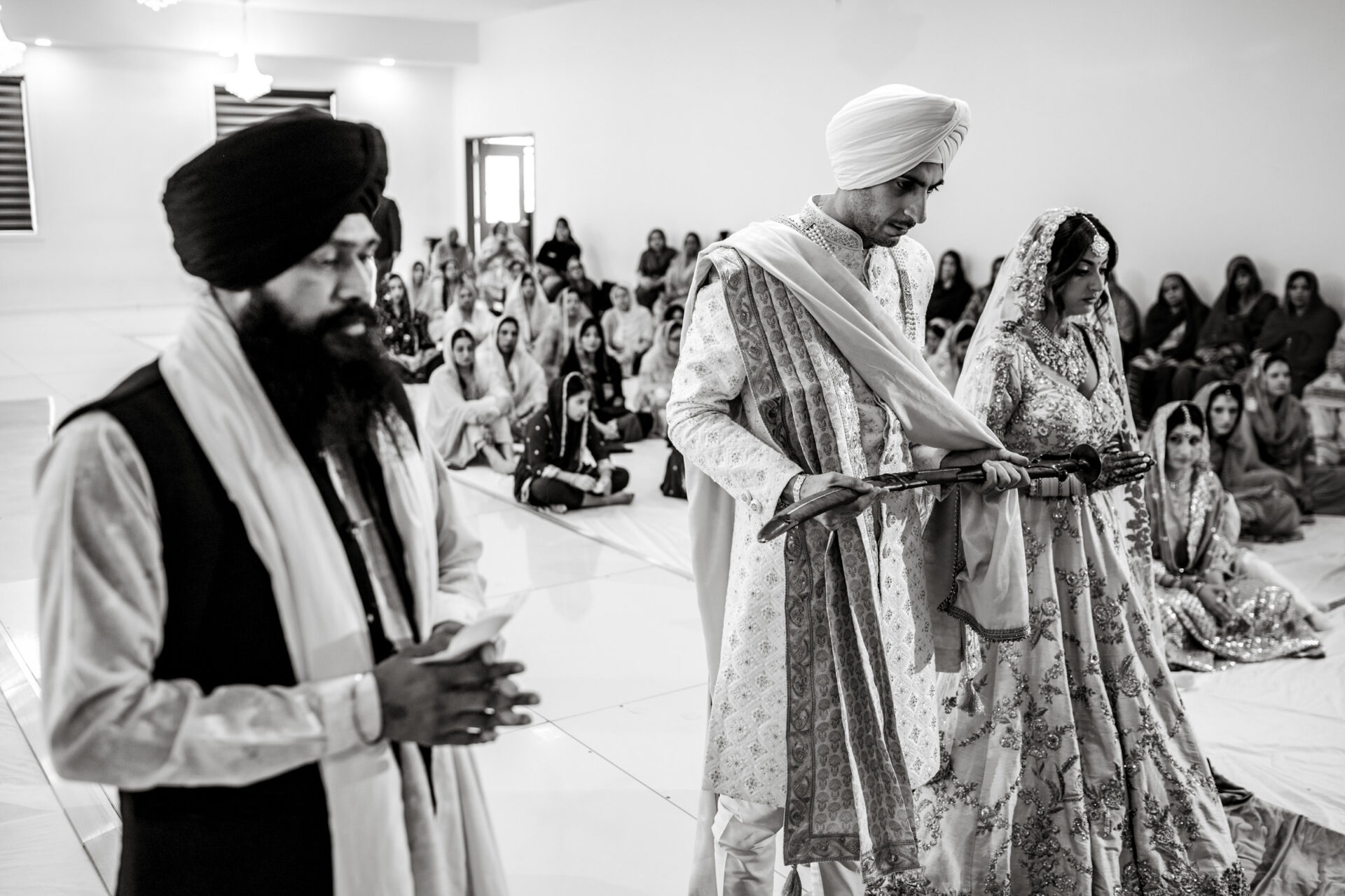 A couple in traditional attire during a Sikh ceremony, guests seated behind at the Hindu Wedding.