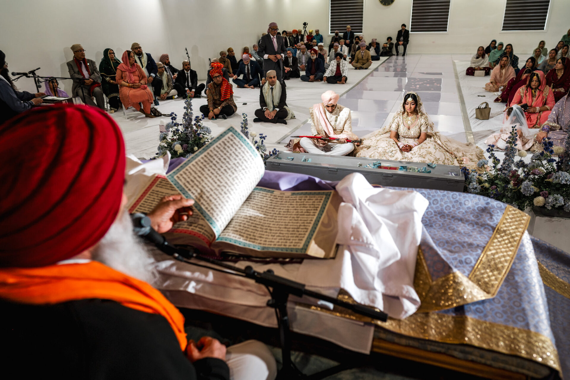 Sikh & Hindu wedding ceremony with a couple seated, officiant reading, guests around.