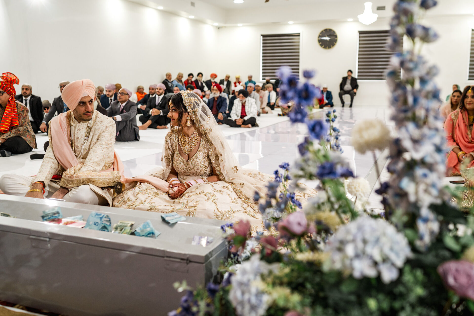 A couple in traditional attire sits in a decorated hall during a Sikh wedding ceremony.