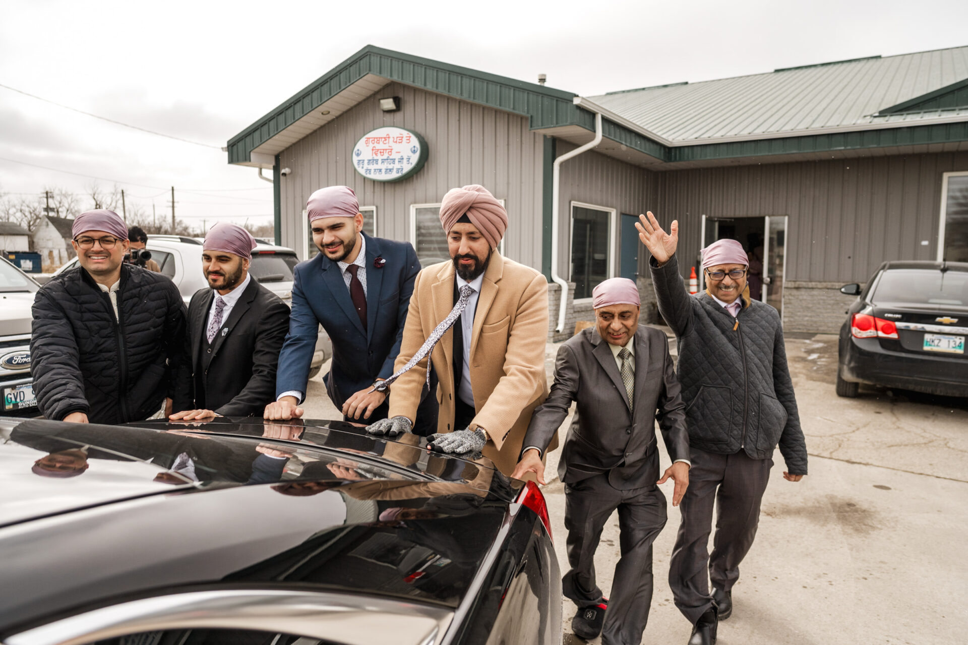 Six men in turbans push a black car, part of a lively Sikh wedding scene on a cloudy day.