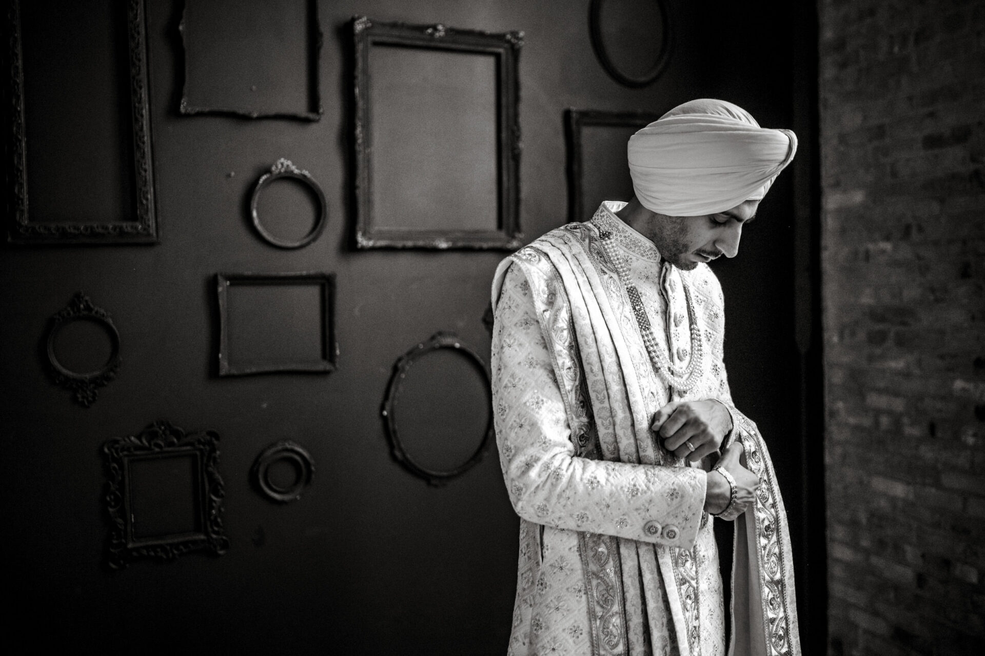 Sikh man in traditional attire adjusts sleeve by empty frames on a dark wall.