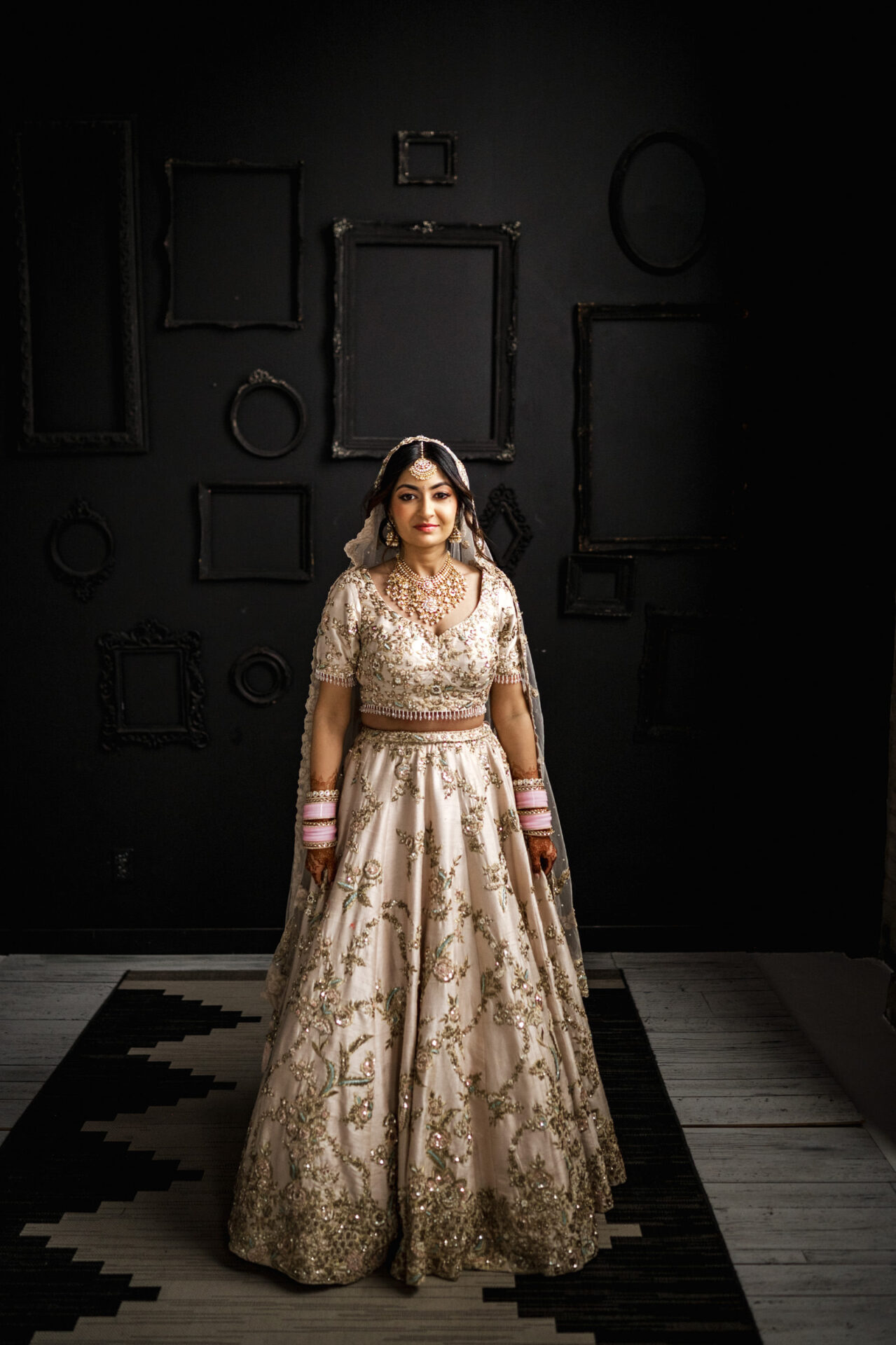 Bride in embroidered dress, standing indoors at a Sikh wedding with empty picture frames.