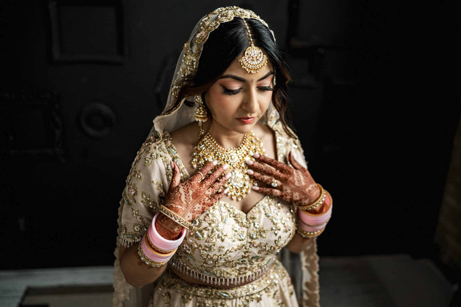 Bride in ornate attire, hennaed hands, and jewelry at a Sikh wedding, softly lit in darkness.