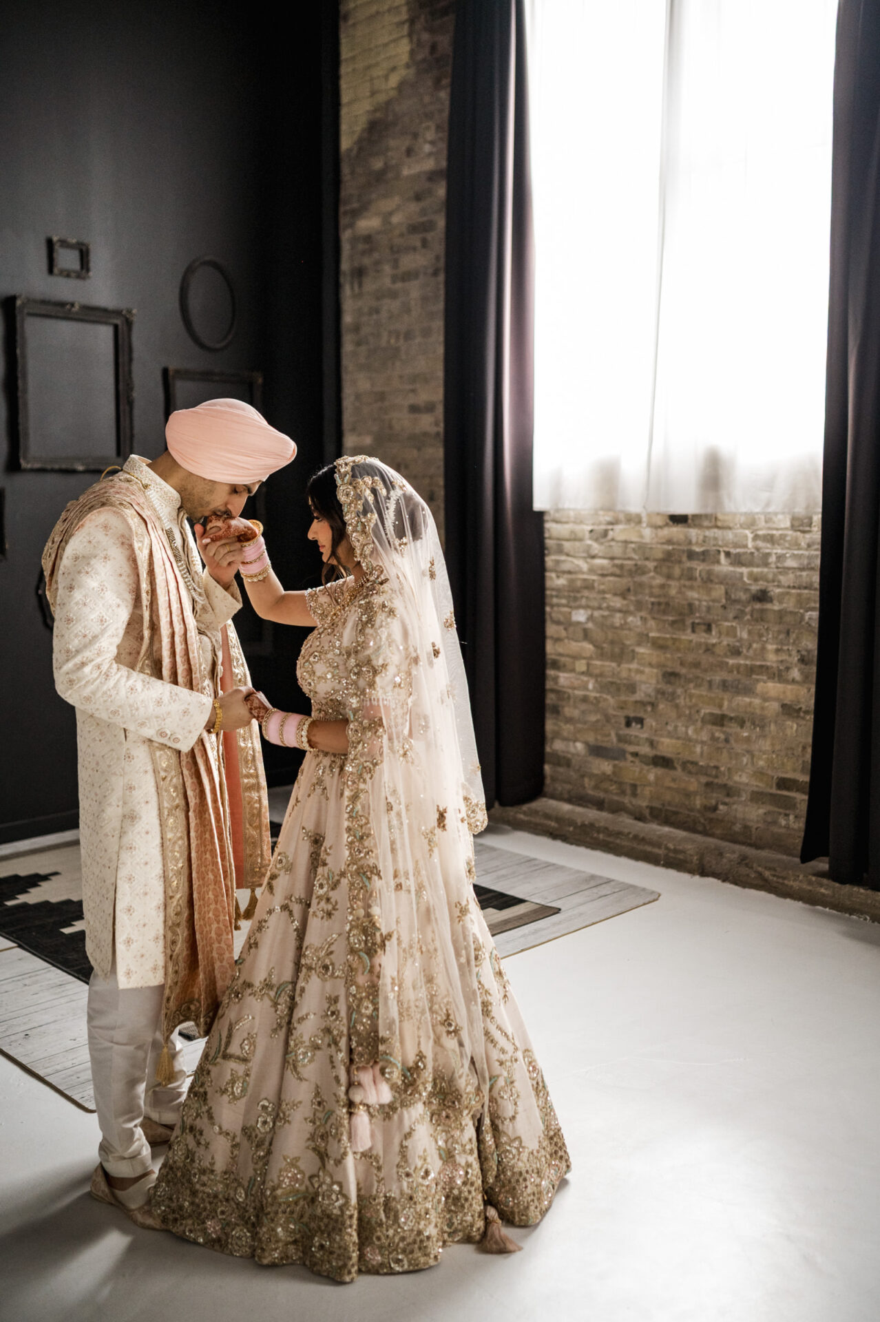 A Sikh couple in traditional attire shares a tender moment in a sunlit room with brick walls.