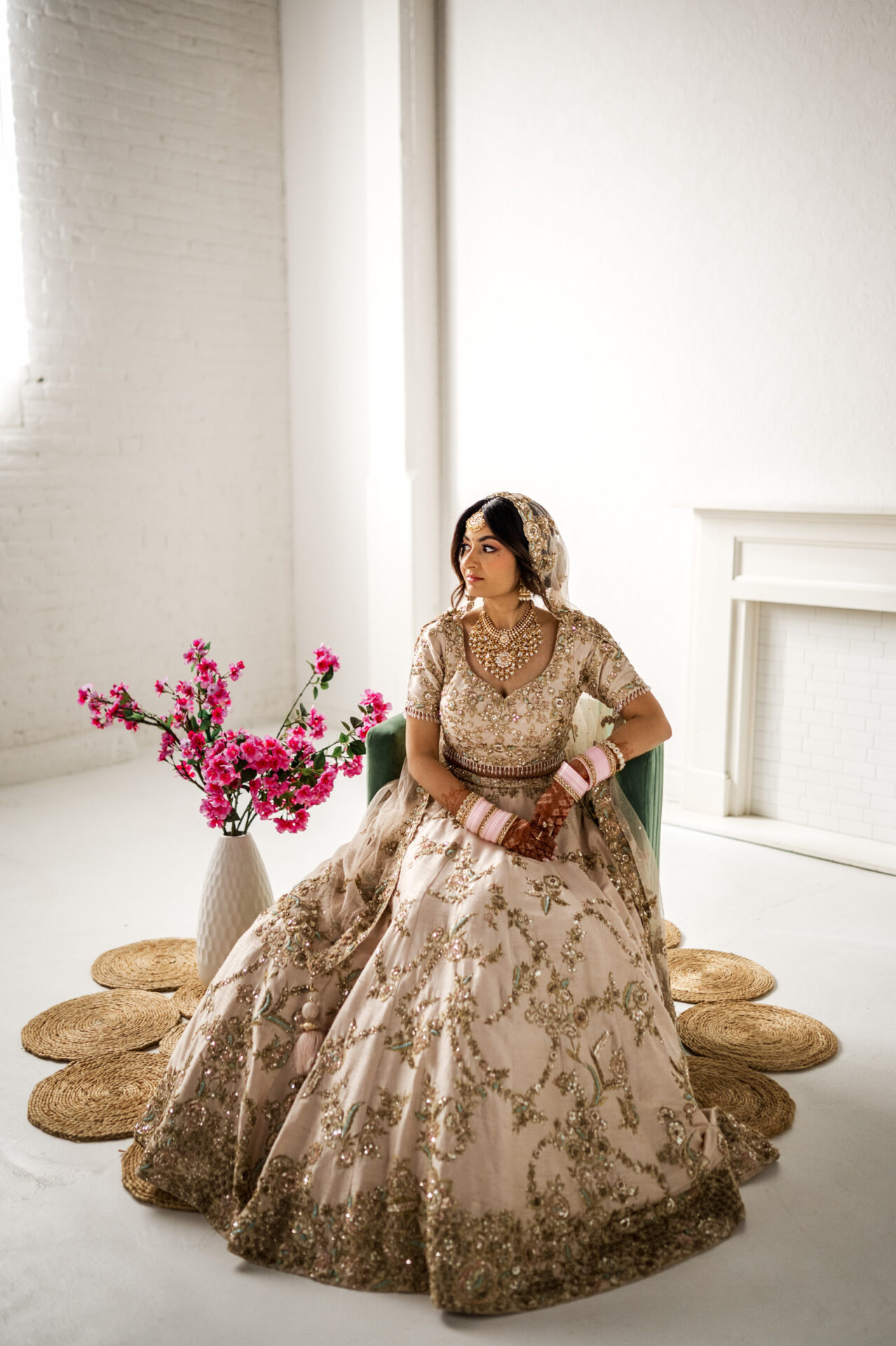 Bride in ornate dress and jewelry sits on chair with pink flowers at Sikh wedding.