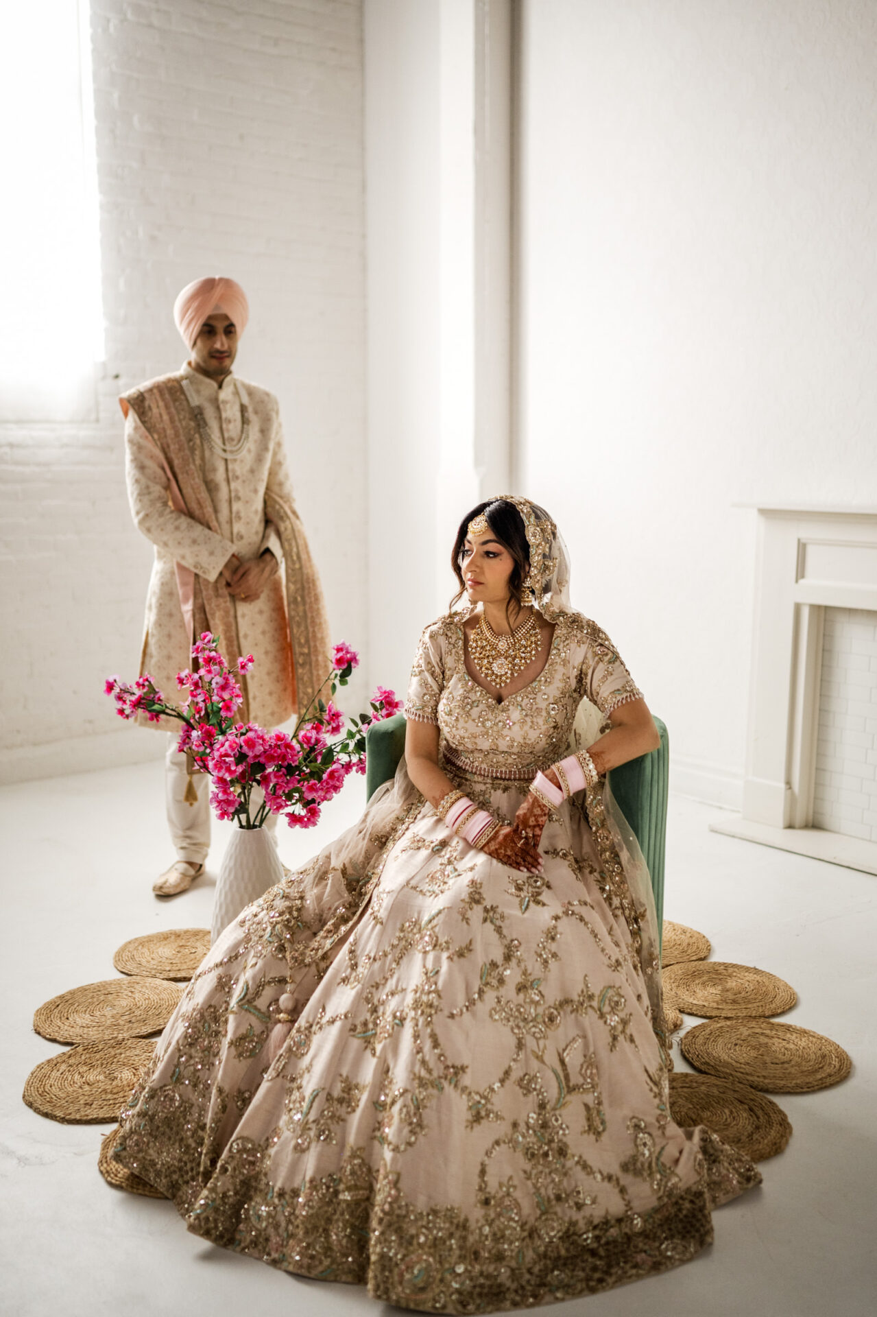 Sikh couple in traditional attire; woman seated, man with flowers in airy, bright room.