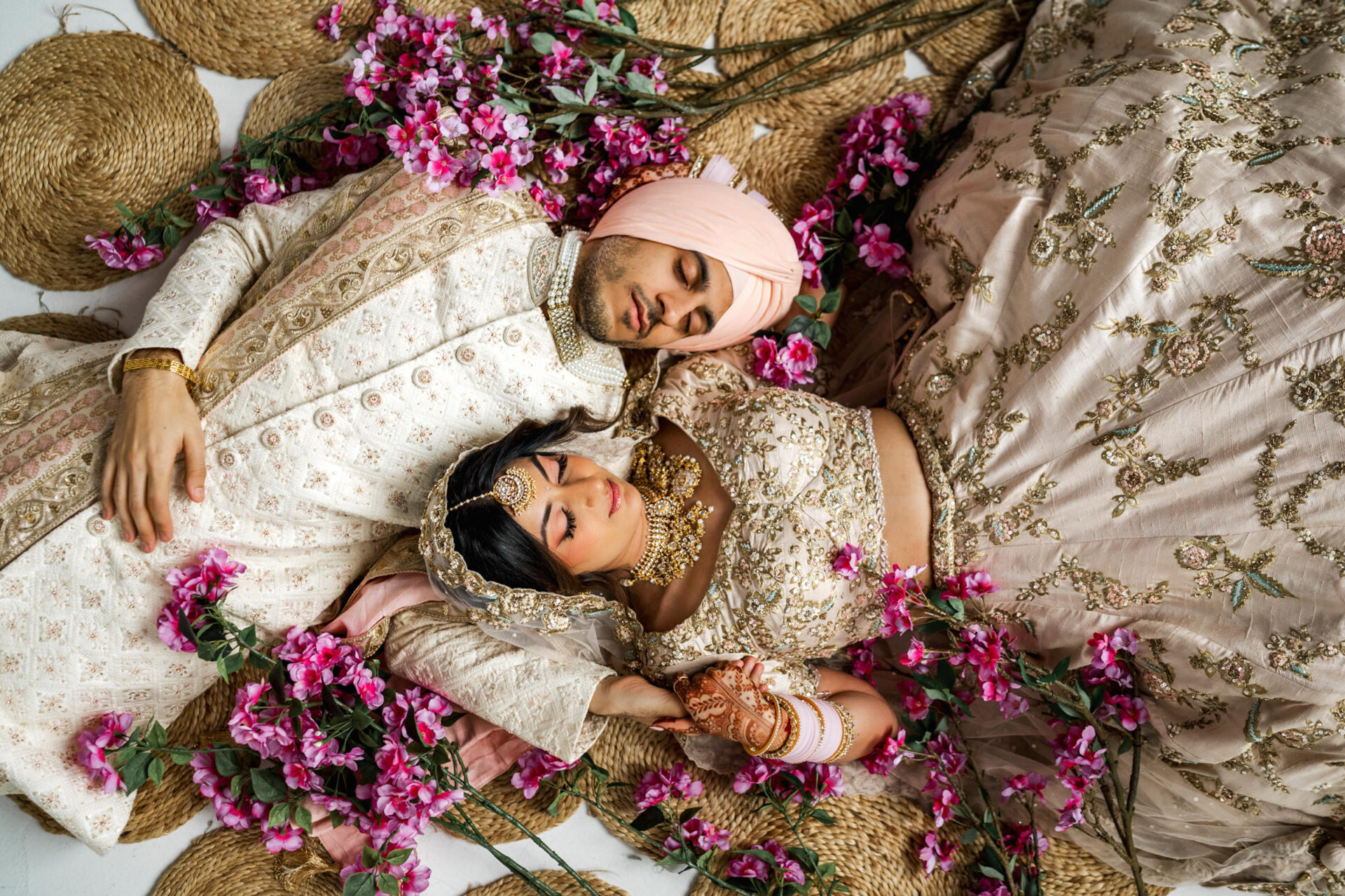 Couple in elegant traditional attire at a Sikh wedding, lying on a bed surrounded by pink blossoms.