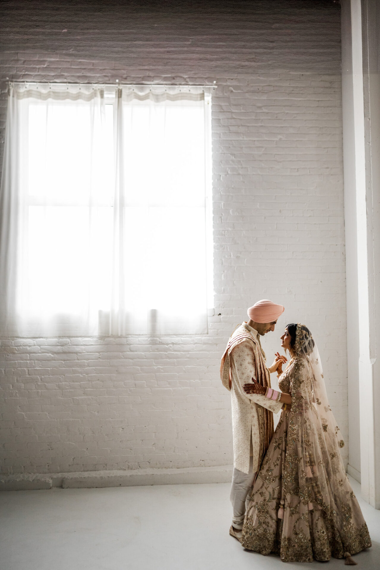 A couple in traditional attire stands by a bright window at a Sikh wedding.