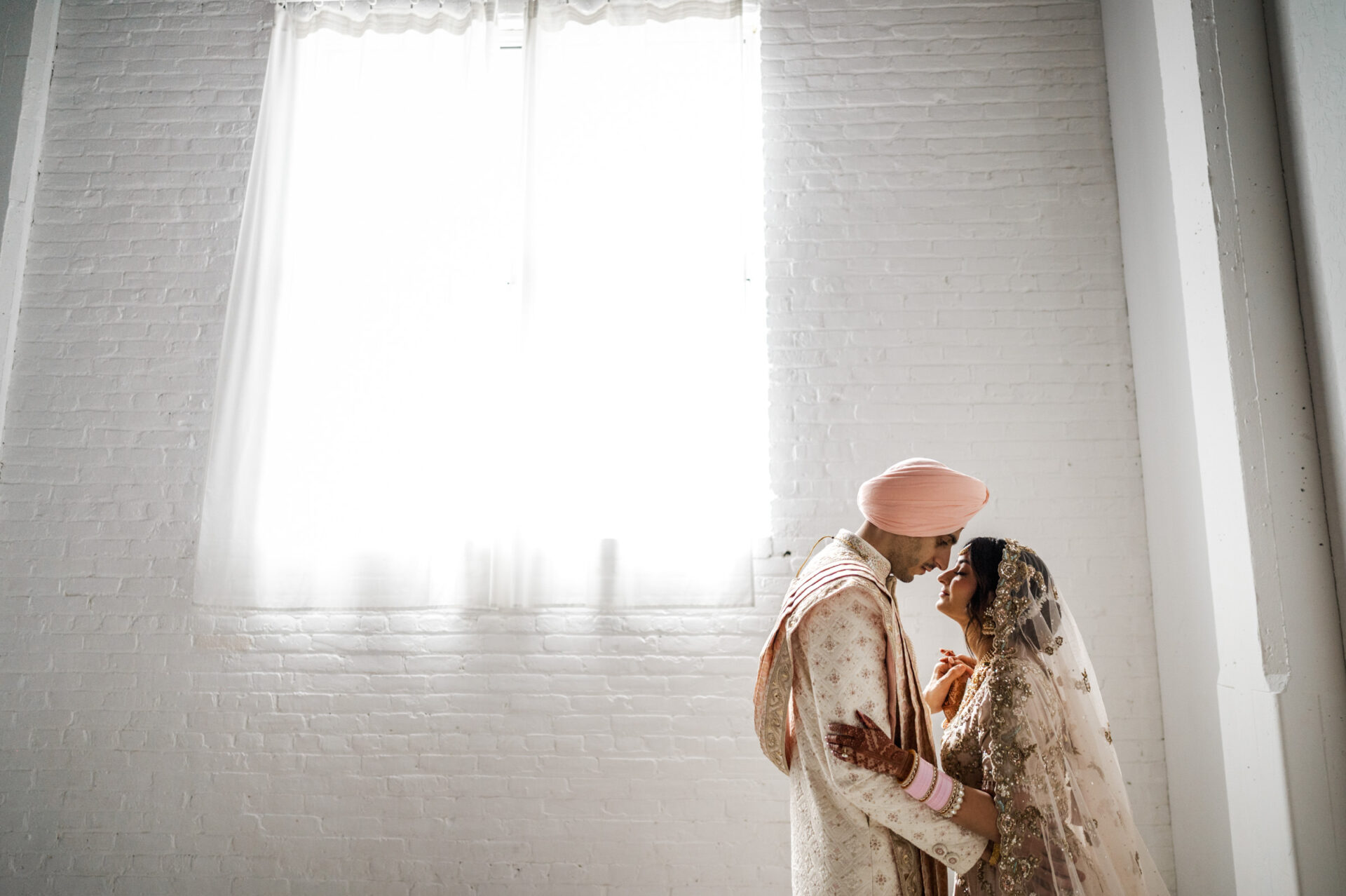 A couple in wedding attire, embracing in a bright, white room at their Sikh Wedding.