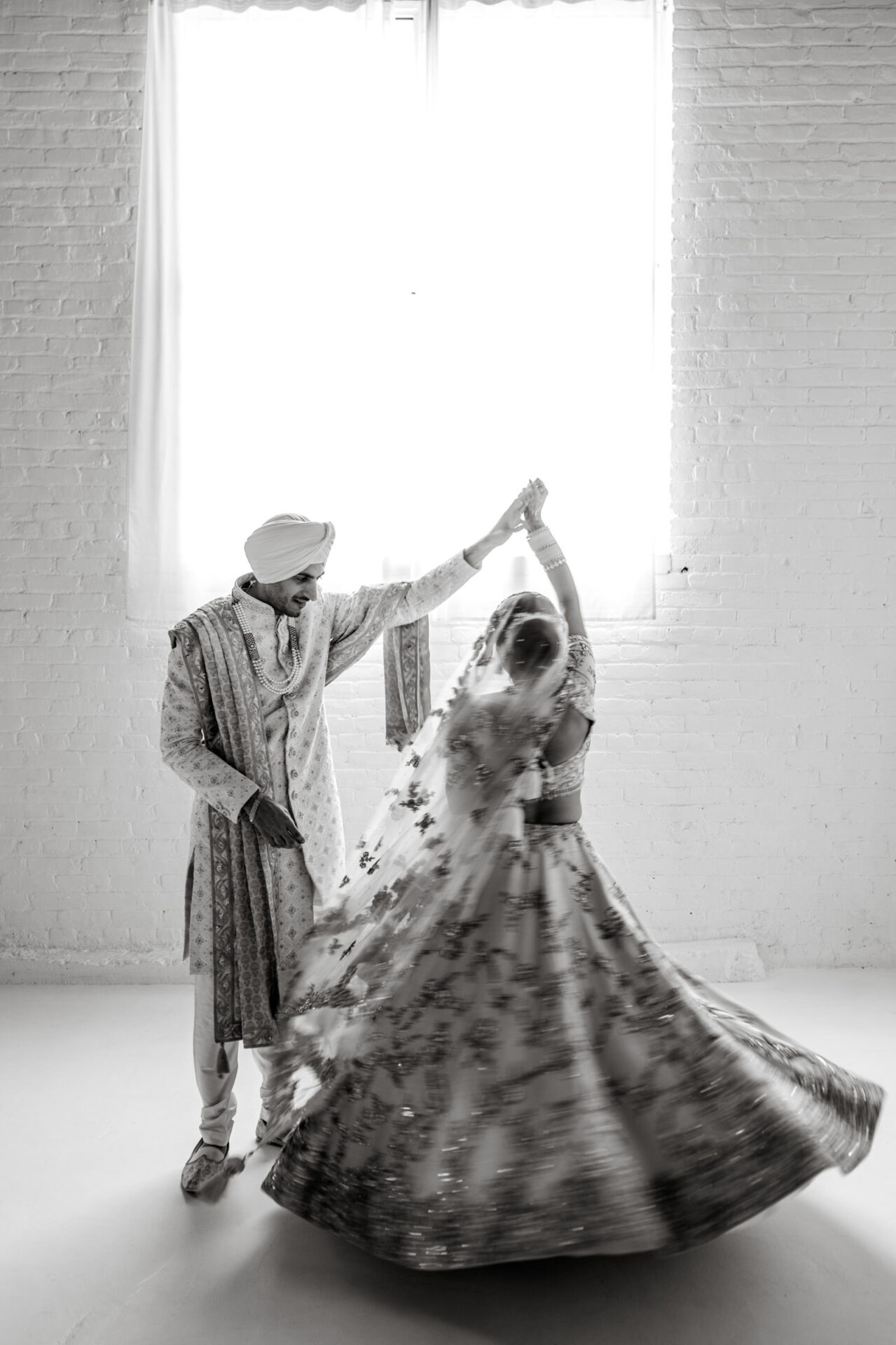 Couple in traditional attire dance at Sikh wedding, swirling dress near bright window.