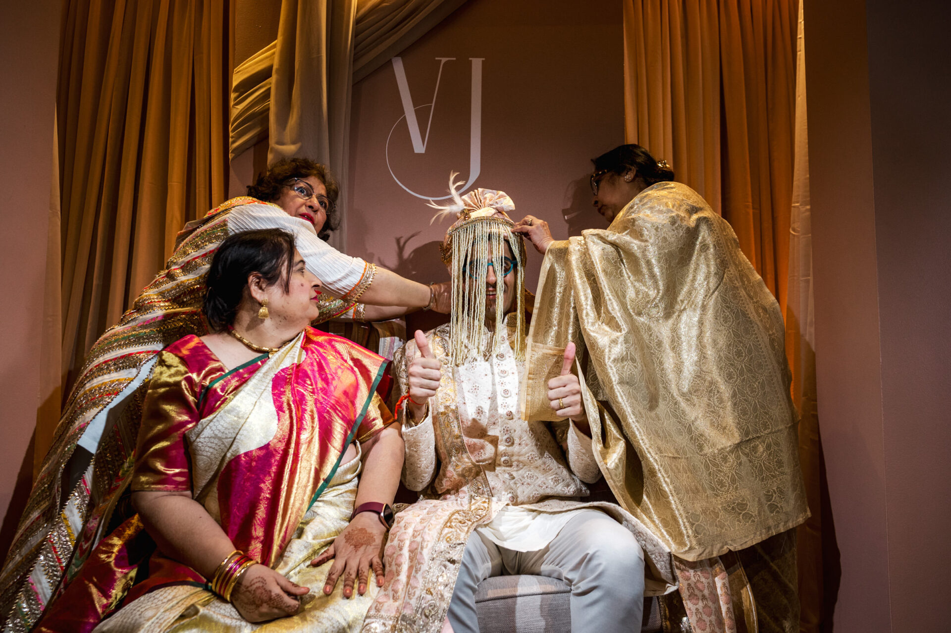 At a Sikh wedding, a groom gives thumbs up as women arrange his attire.
