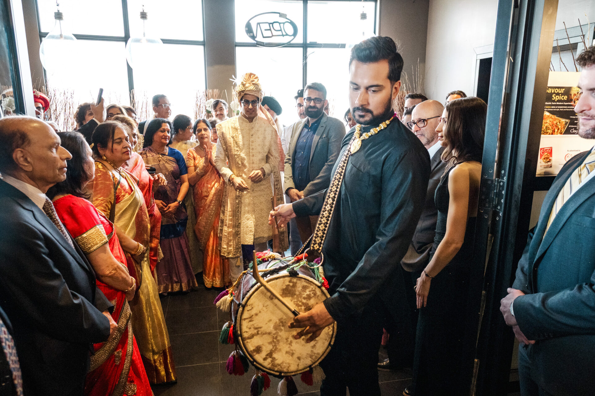 A drummer leads a lively Sikh wedding procession with guests in colorful, traditional attire.