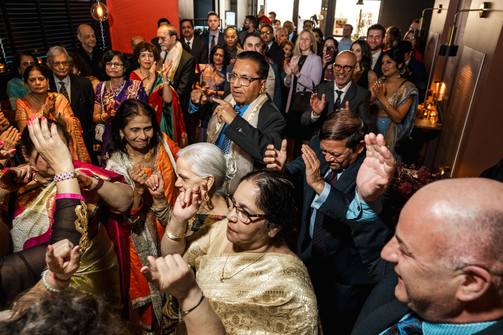 People in formal attire dancing joyfully at a lively indoor Sikh wedding.
