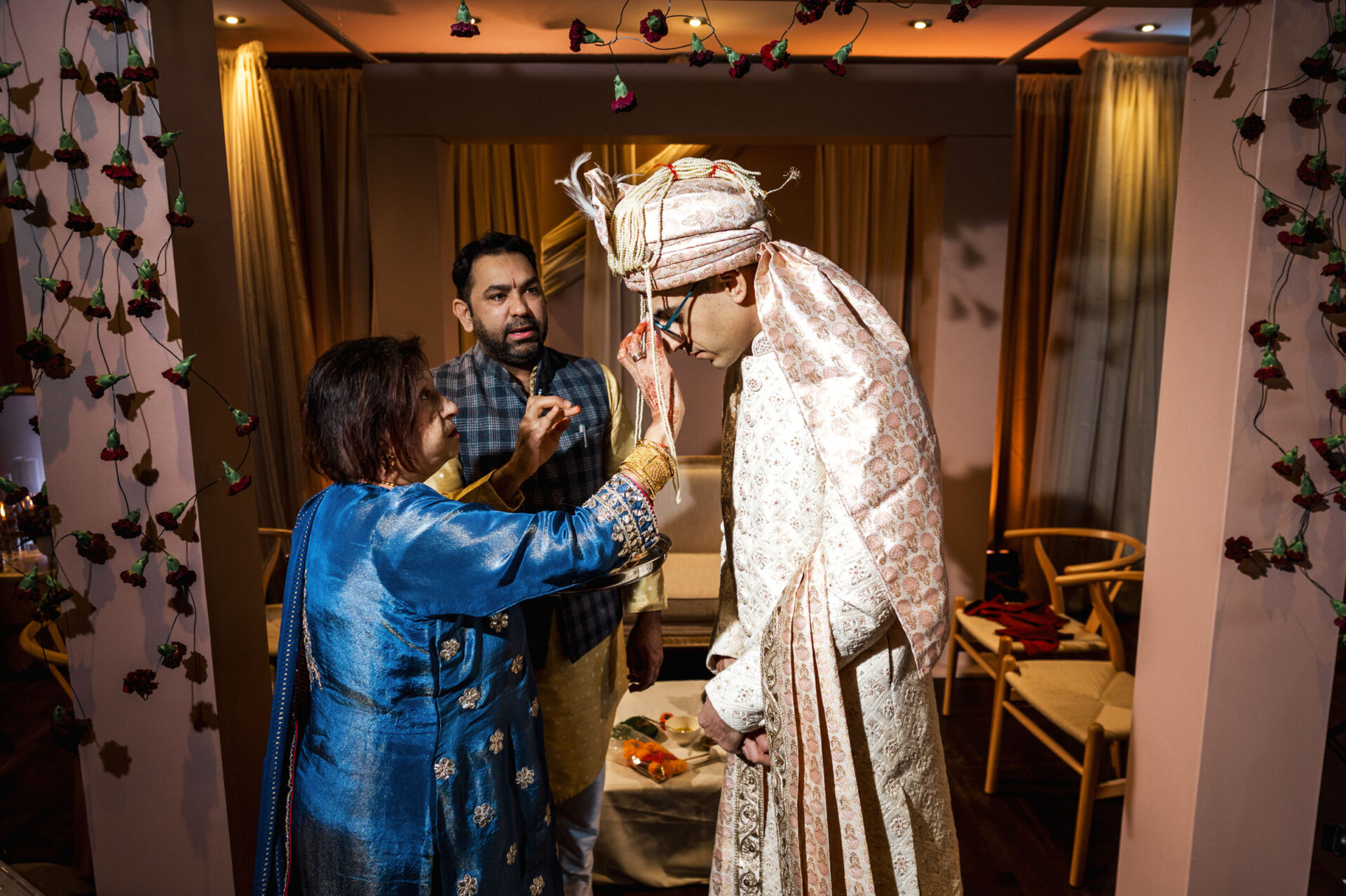 A woman blesses a groom in traditional Sikh attire as another man stands nearby.