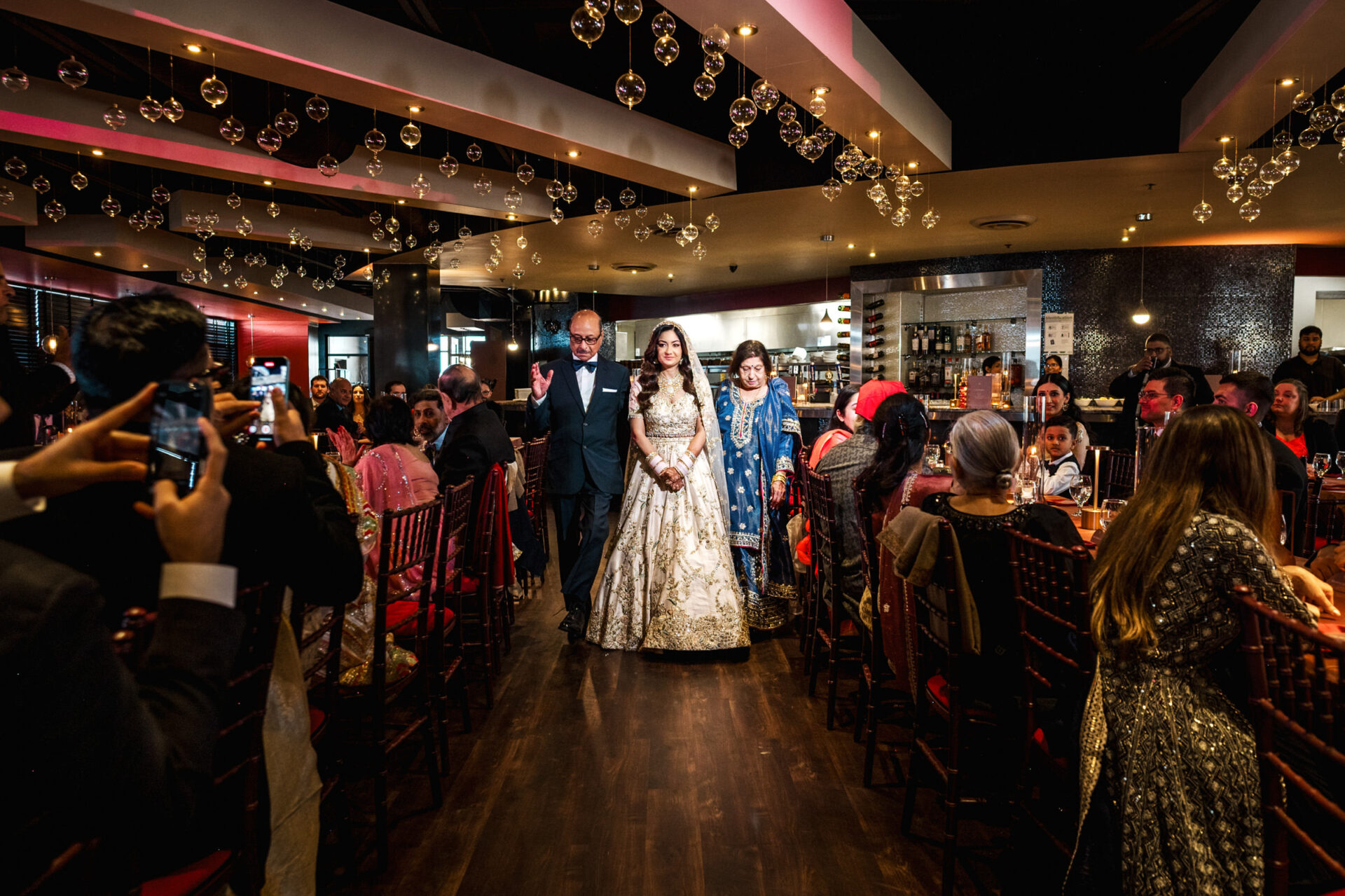 A bride walks with two escorts in a decorated banquet hall during a Sikh ceremony.