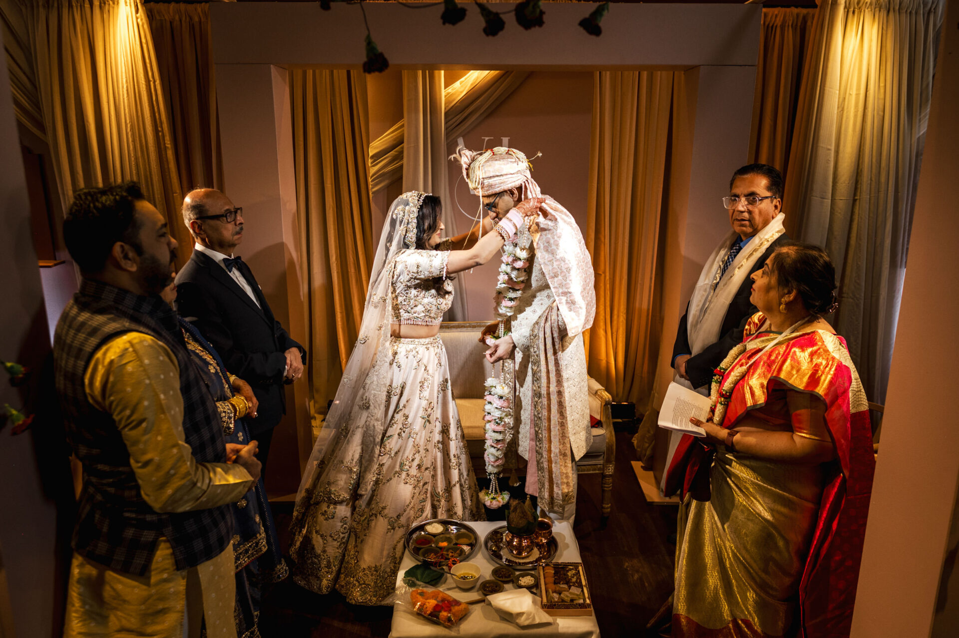 A Sikh couple in traditional attire at an indoor wedding, surrounded by guests.