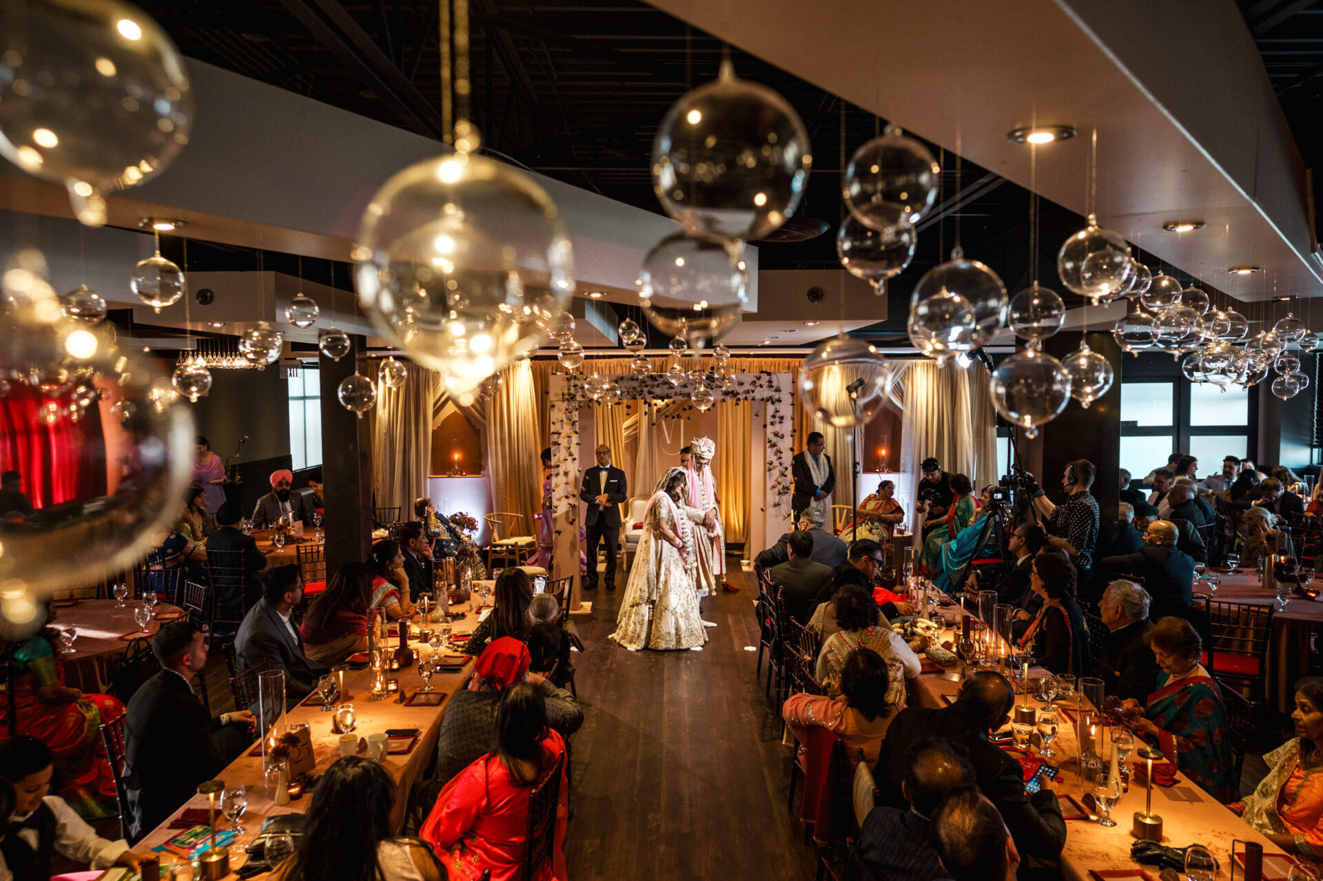 A Sikh wedding ceremony with a decorated venue and guests under hanging glass orbs.