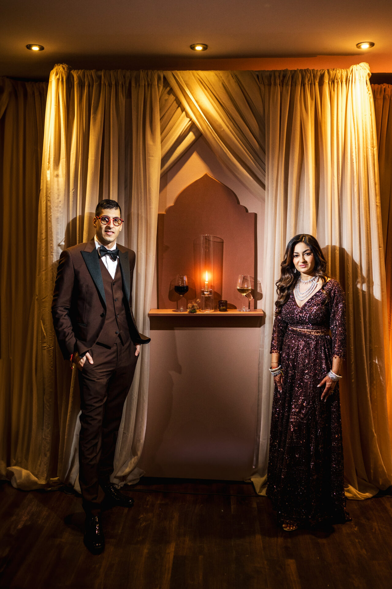 A couple in formal attire, poised at a Sikh & Hindu wedding with soft ambient lighting.