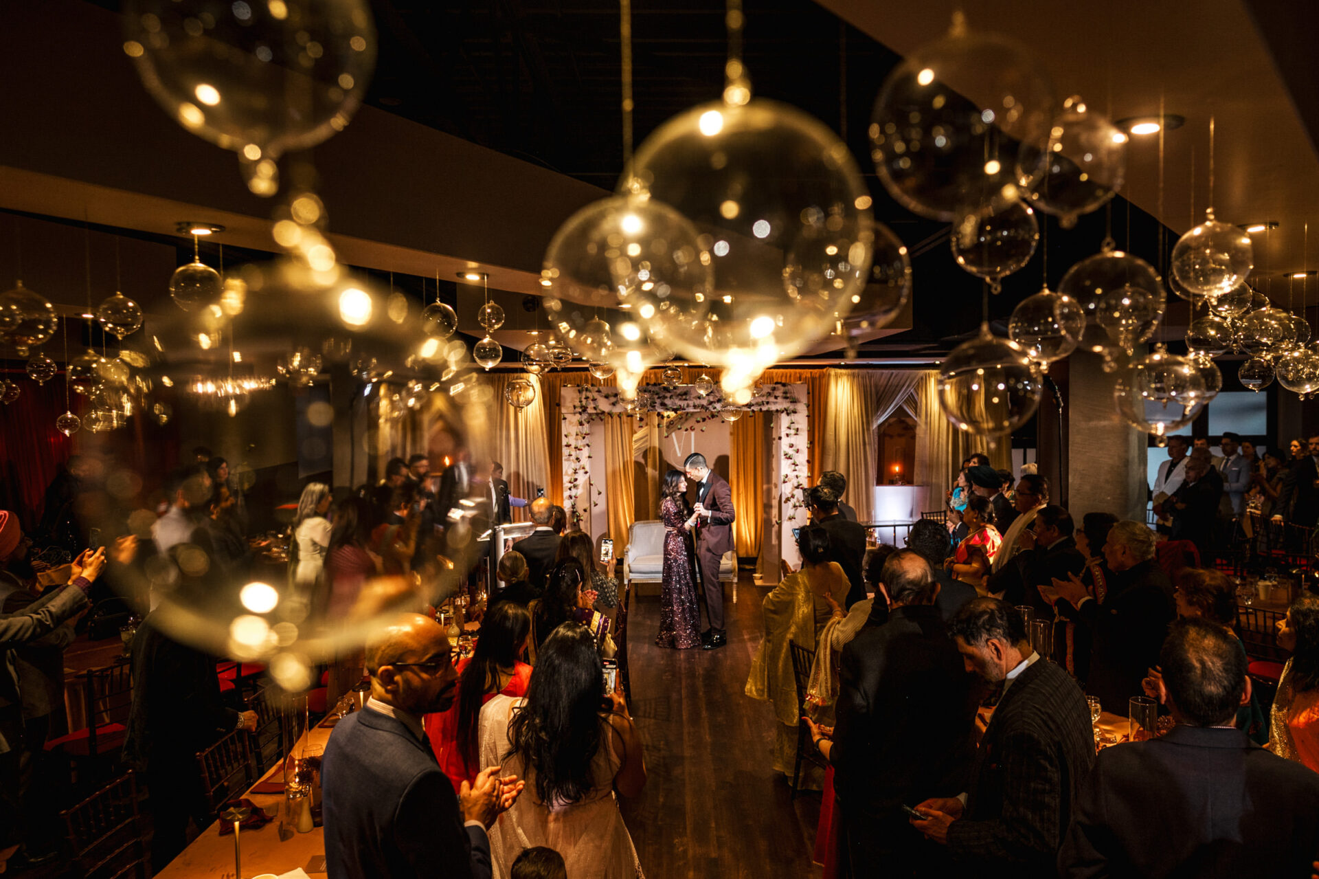 A couple dances at a Sikh wedding, surrounded by guests and hanging glass orbs.