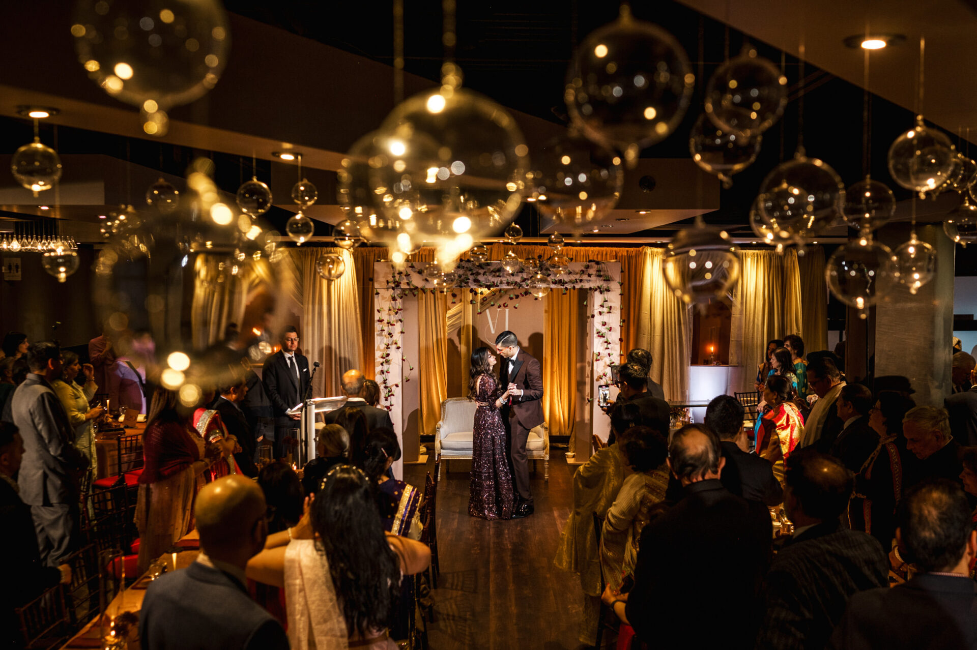 A couple stands on a decorated stage at a vibrant Sikh wedding, surrounded by guests and lights.