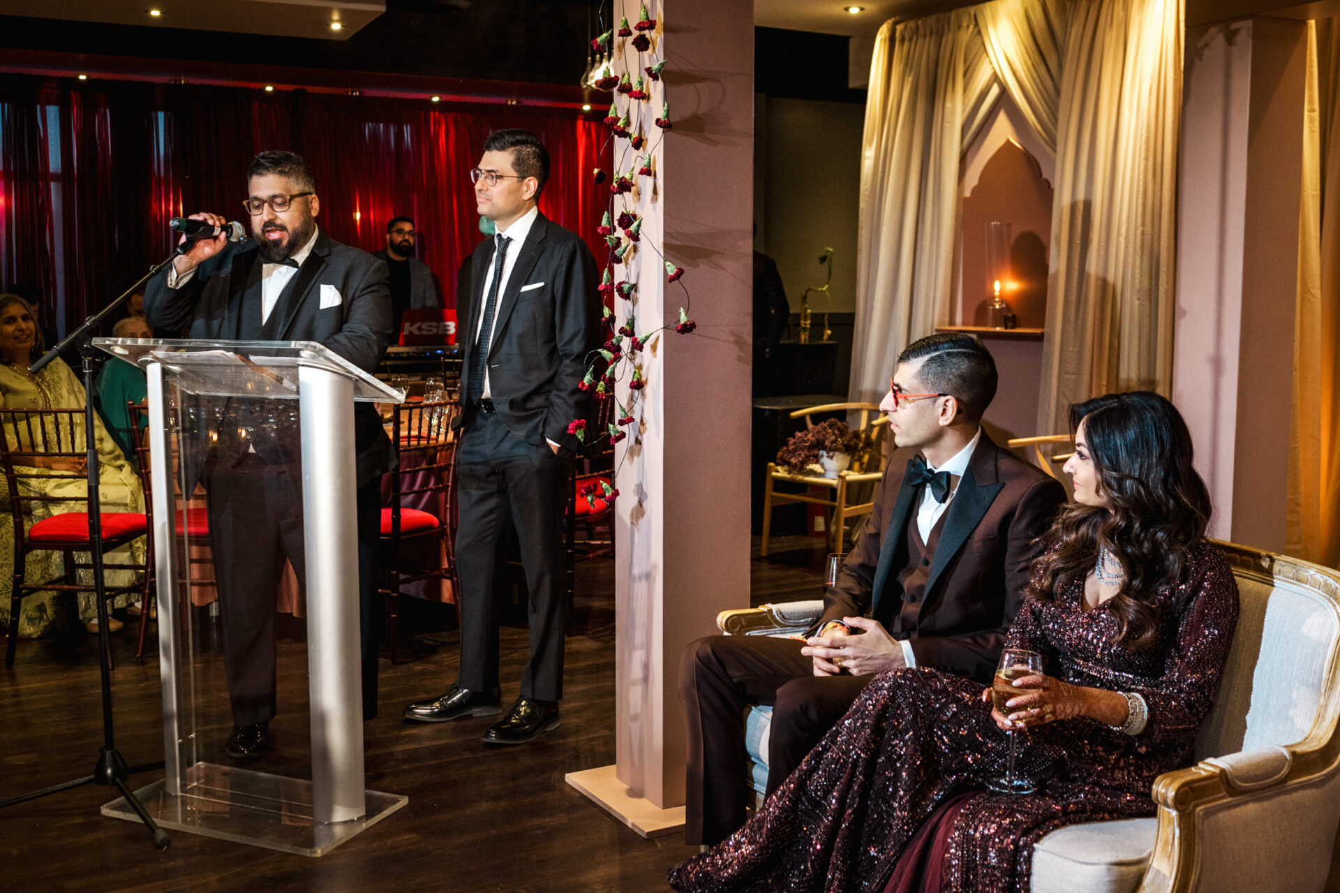 A man speaks at a podium during a Sikh wedding; a couple sits nearby with guests in the background.