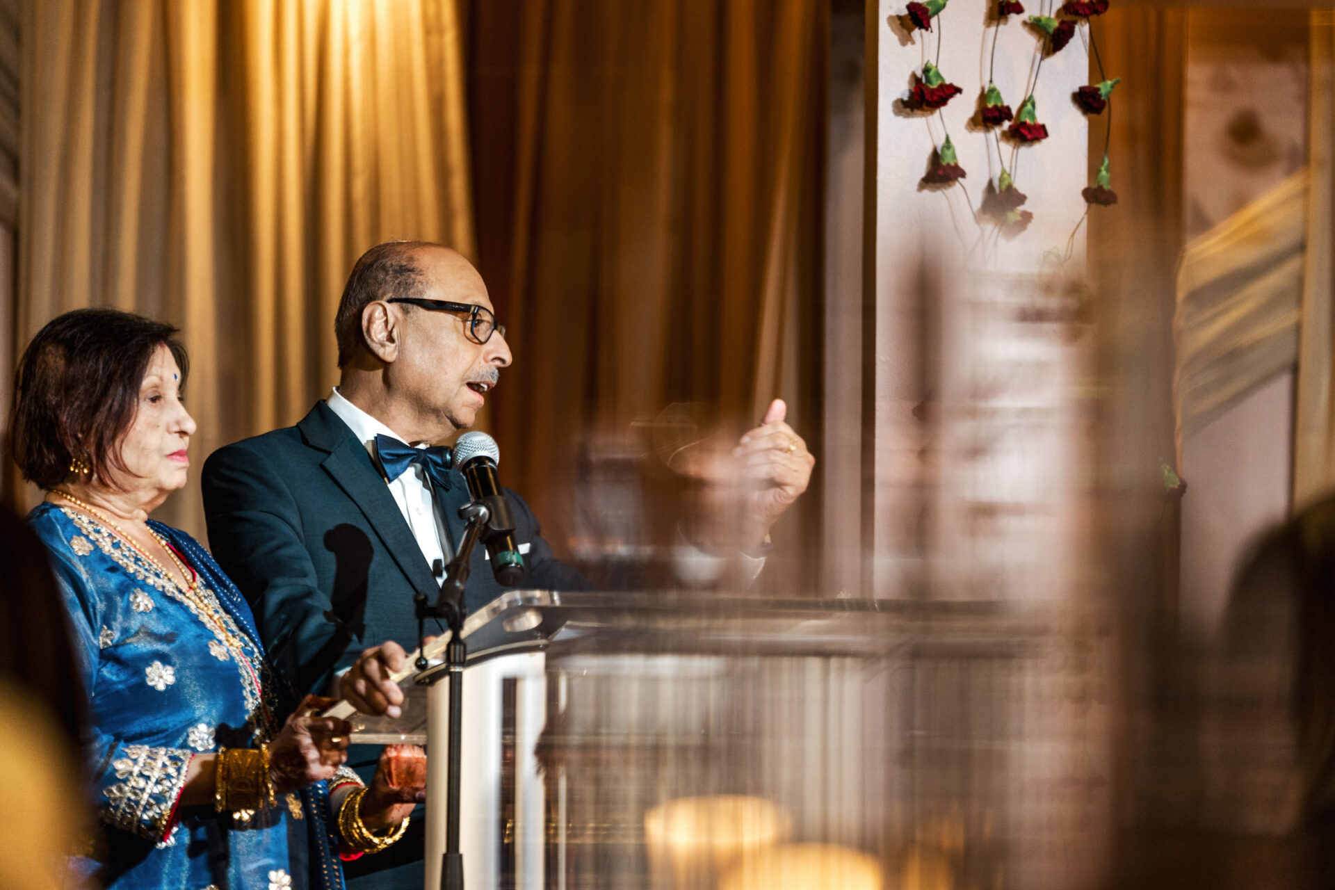 An elderly couple speaks at a podium during a Sikh wedding with warm lighting.