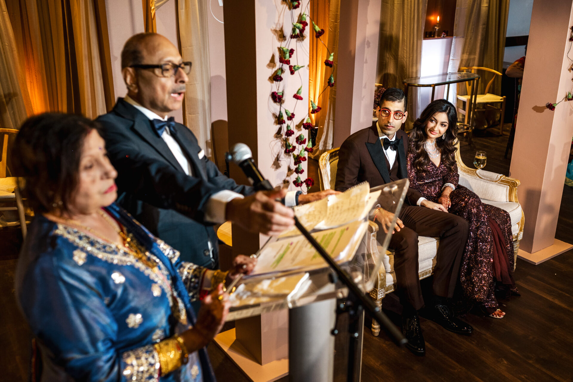 A couple sits as two people speak at a microphone during a formal Sikh wedding.