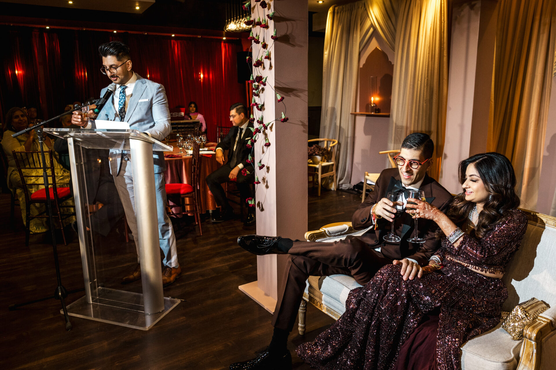 A couple clinks glasses as a man speaks at the podium during an elegant Sikh wedding.