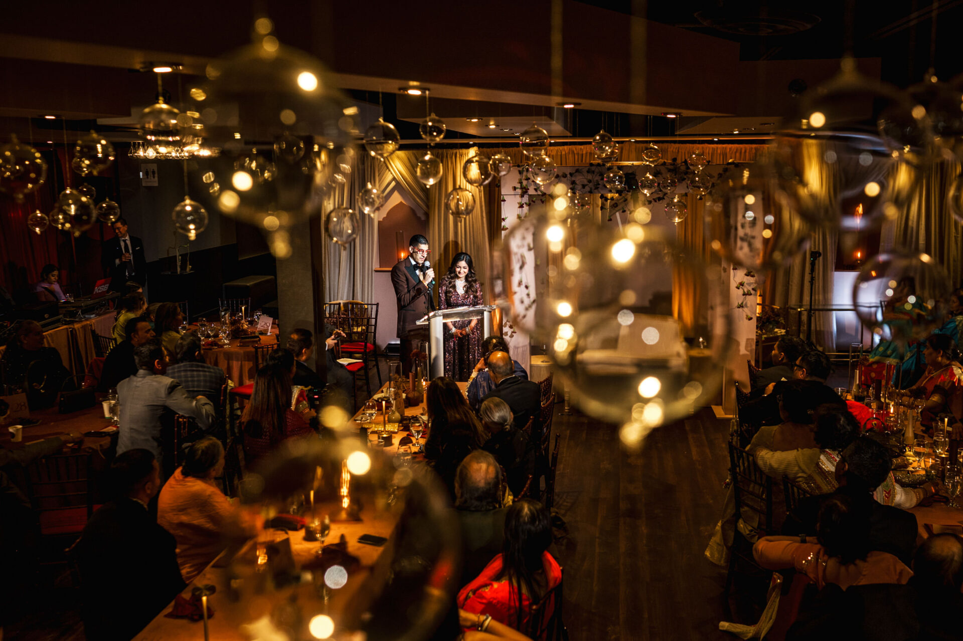 A couple speaks on stage at an elegant Sikh wedding, lights twinkling over the seated audience.