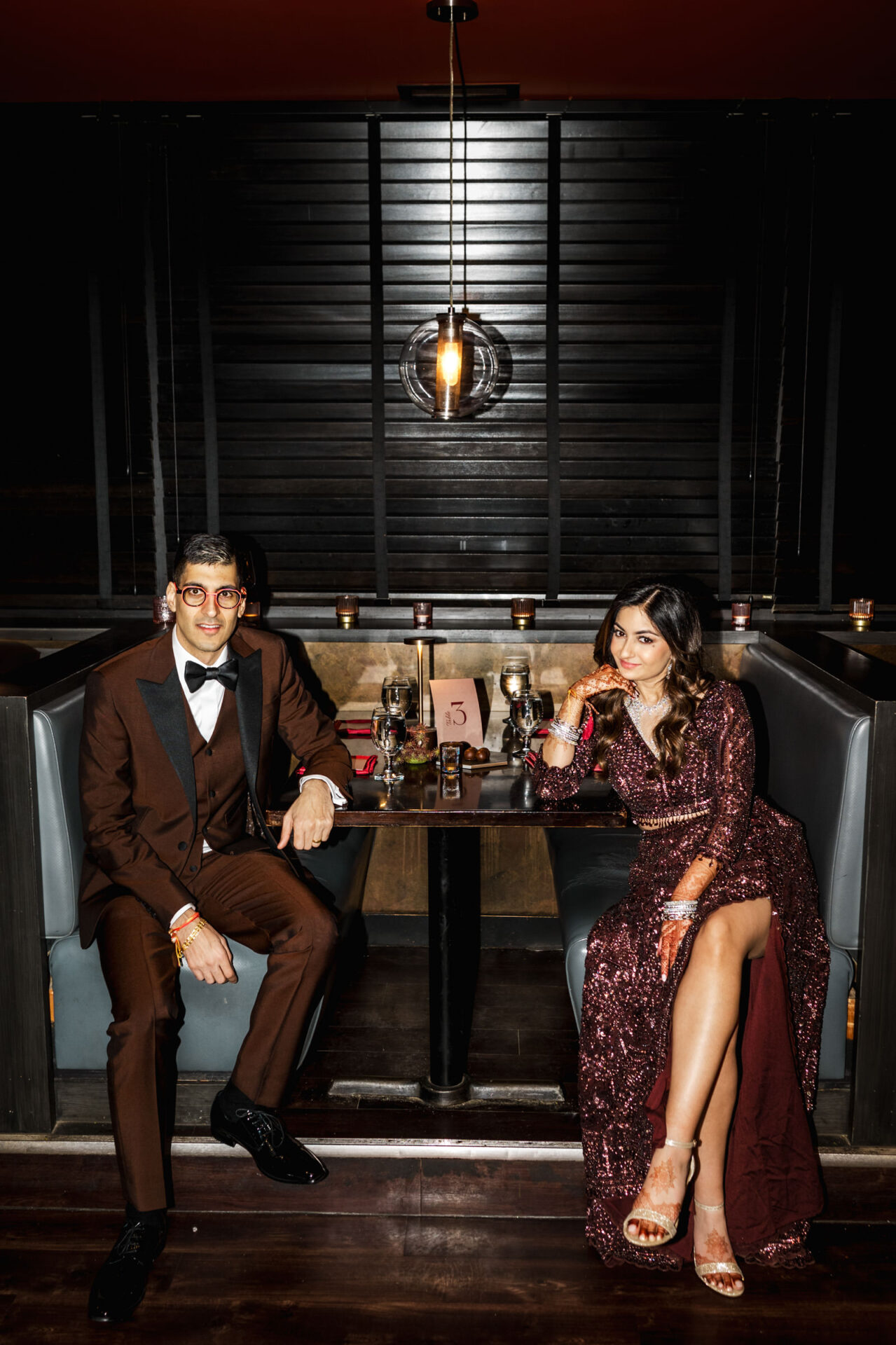 Couple in brown attire at a dimly lit booth, smiling like it's a Sikh wedding moment.