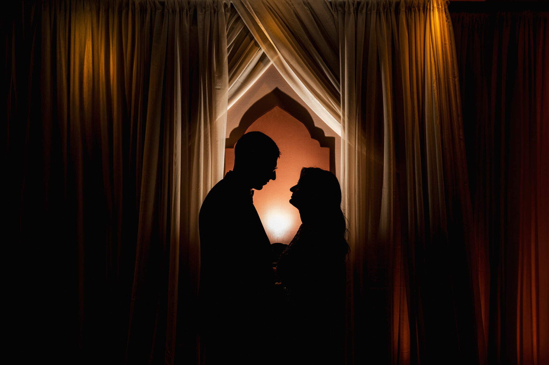Silhouetted couple at a Sikh wedding with a warm, glowing backdrop and draped curtains.