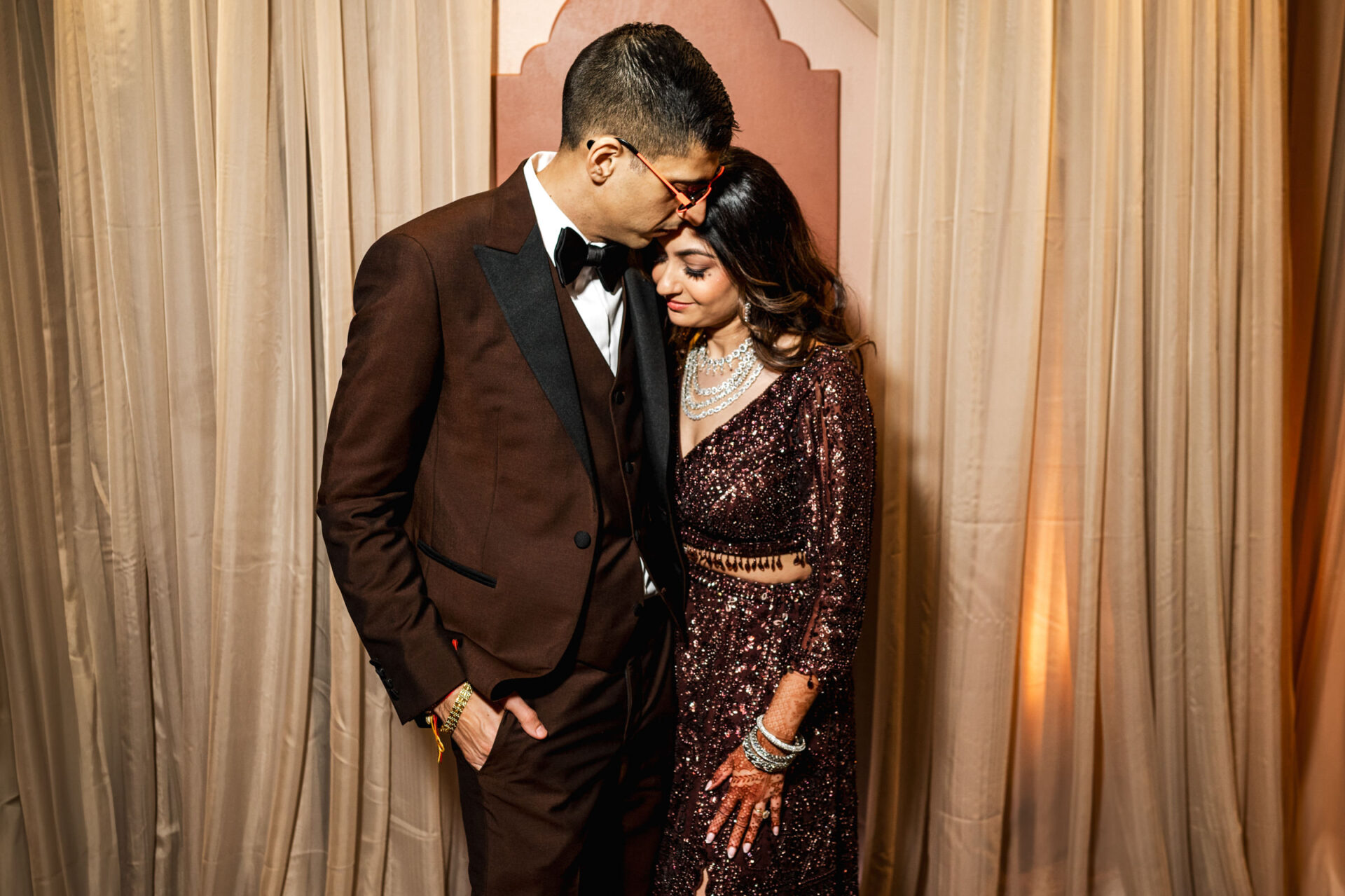 A couple in formal attire embraces, echoing a Sikh wedding vibe by elegant curtains.