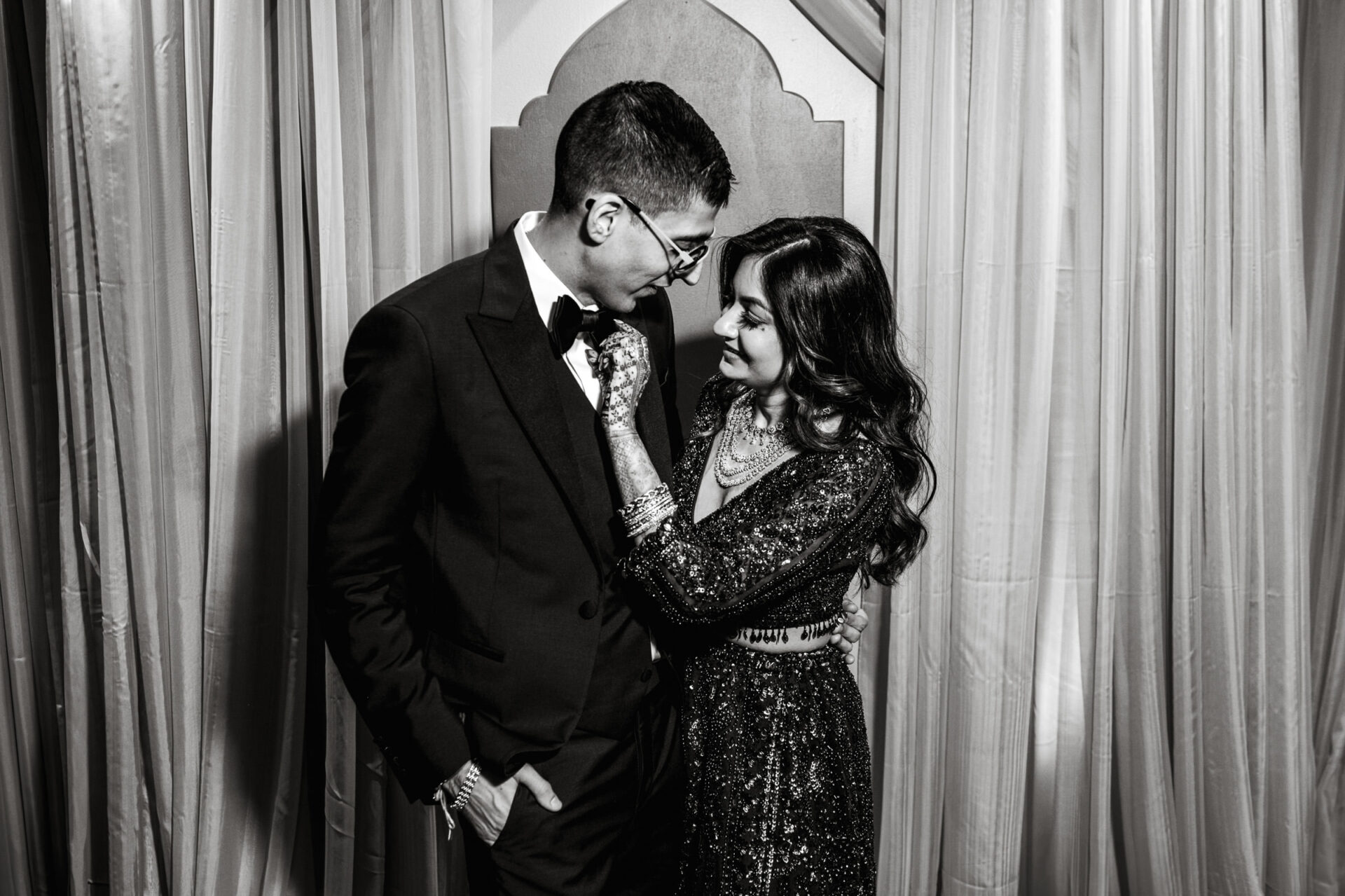 Couple in formal attire embraces at a Hindu wedding, smiling before elegant drapes.