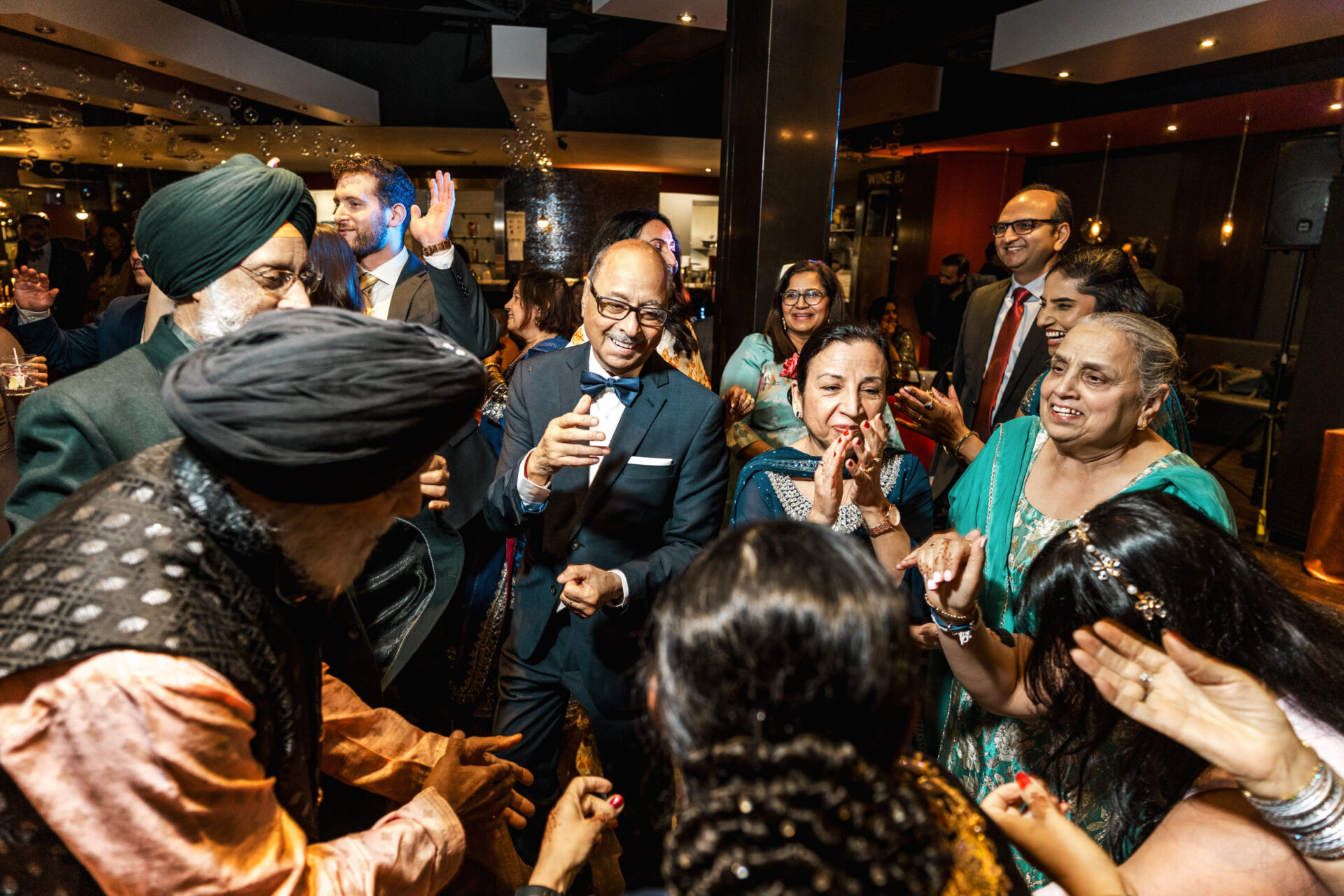 People joyfully dancing at a lively indoor Sikh and Hindu wedding celebration.