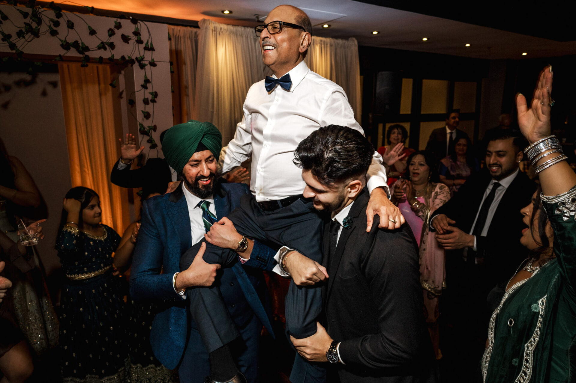 Two men joyfully lift an elder in a Sikh wedding celebration.