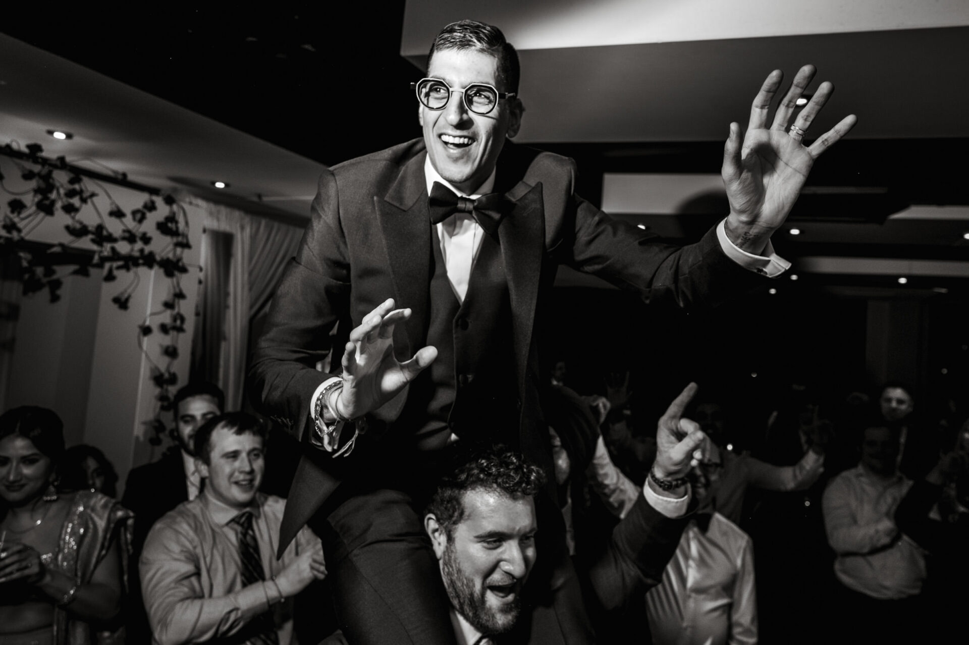 Man in a suit joyfully lifted, celebrating at a lively Sikh wedding.