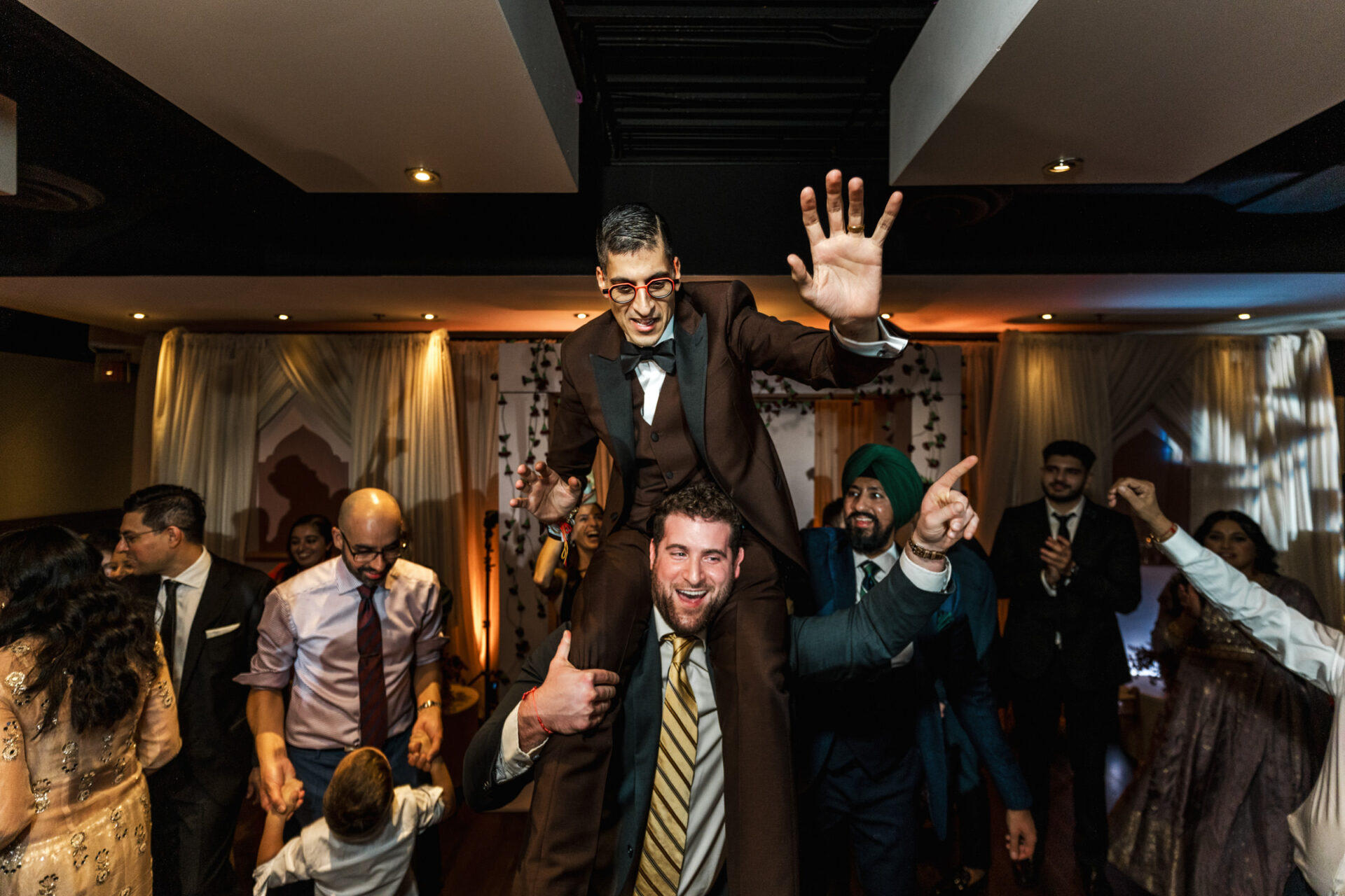 Man in brown suit lifted at lively Sikh wedding, smiling and waving amid dancing people.