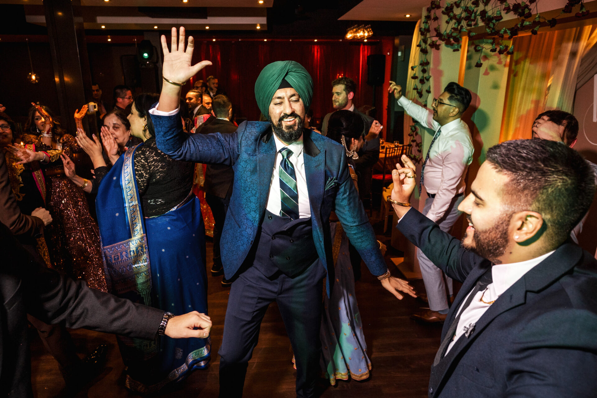 Man in a blue suit joyfully dancing at a lively Sikh wedding party.
