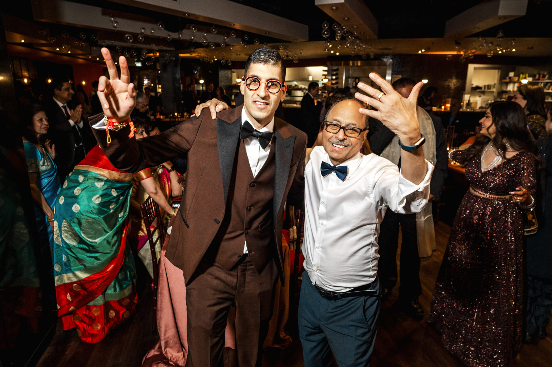Two men smiling at a lively Sikh wedding with people dancing in the background.