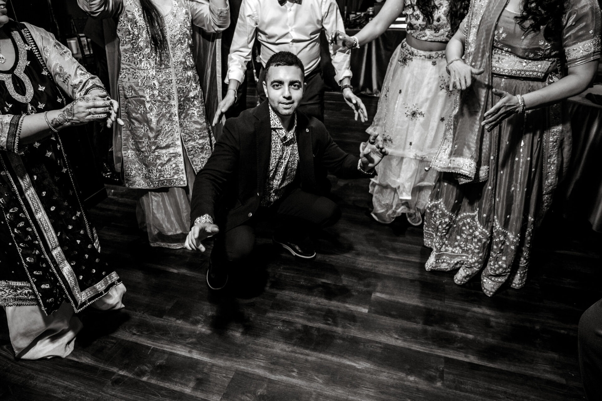 Person crouching on dance floor at a vibrant Sikh wedding, surrounded by traditional attire.