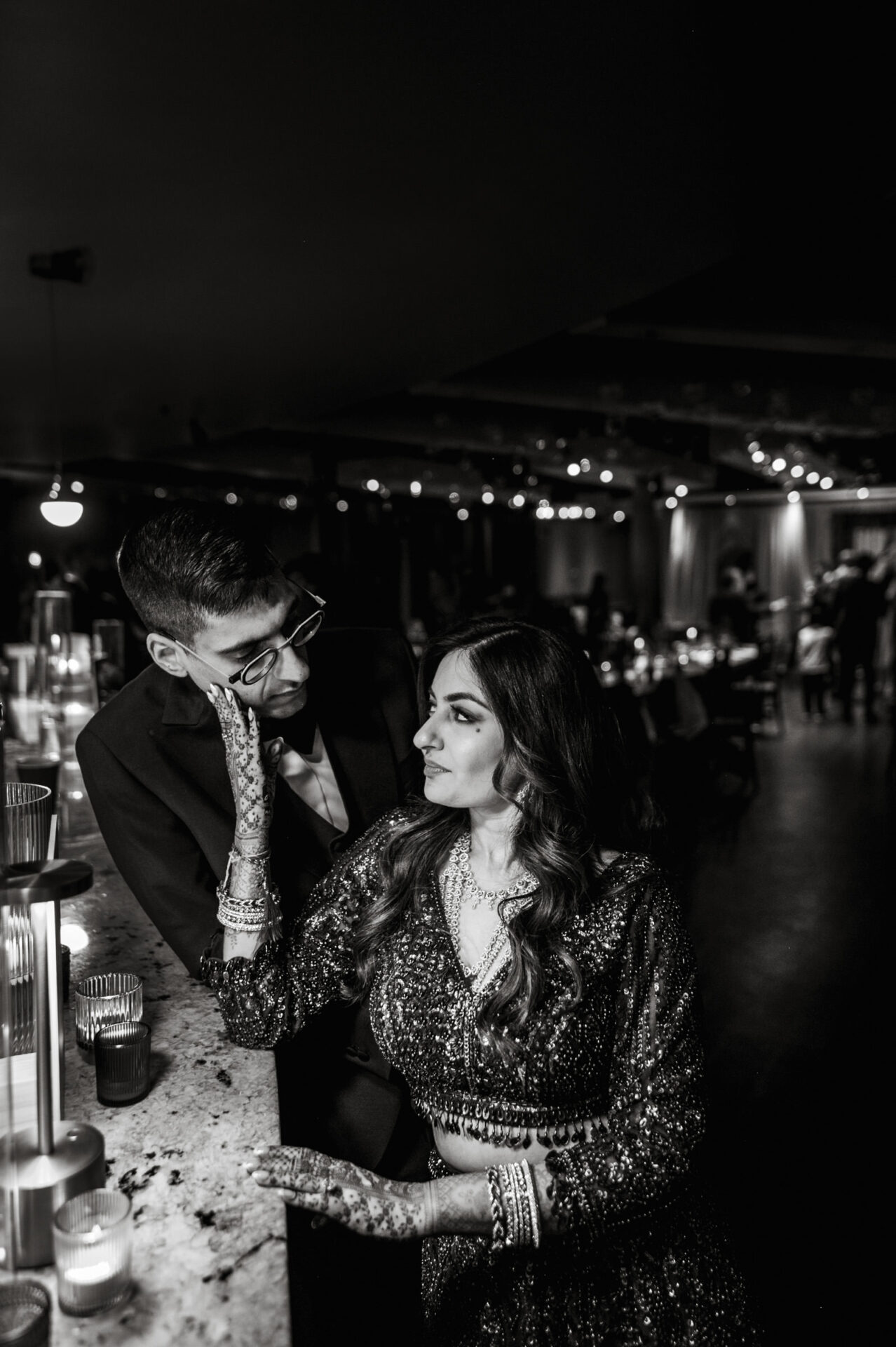 A couple in formal attire shares a gaze at a Sikh wedding in a dimly lit room.