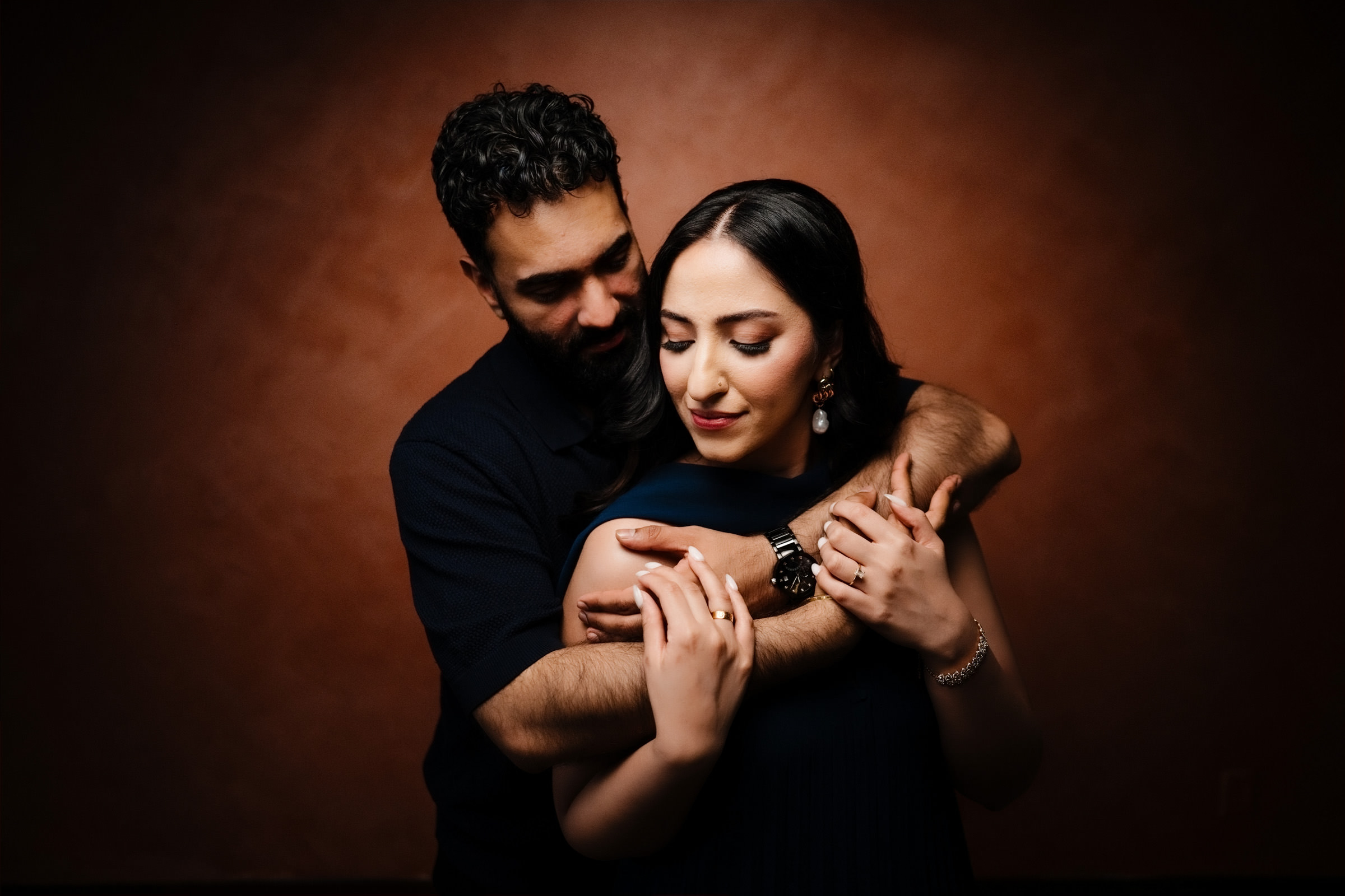 Studio portrait of engaged couple in navy outfits cuddling against a warm brown backdrop